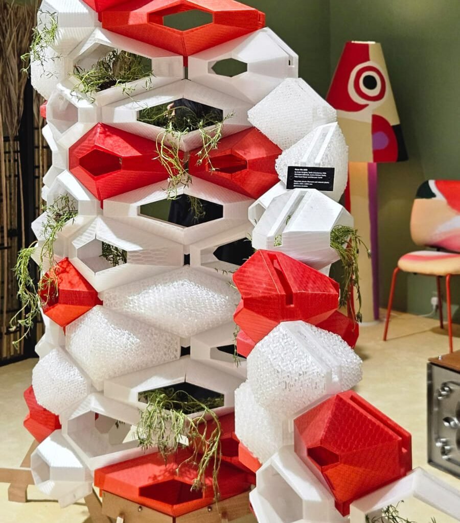 Uma divisória modular que une impressão 3D e biofilia. A estrutura geométrica, em vermelho e branco, funciona como uma colmeia high-tech, integrando plantas ao design. Uma solução funcional e escultural para dividir espaços.