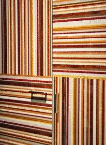Marcenaria como obra de arte. Este armário usa lâminas de madeira em tons quentes (vermelho, amarelo, marrom) para criar um padrão gráfico de listras. A alternância entre horizontal e vertical, combinada com puxadores modernos, resulta em um design arrojado e luxuoso.