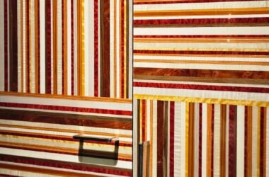 Marcenaria como obra de arte. Este armário usa lâminas de madeira em tons quentes (vermelho, amarelo, marrom) para criar um padrão gráfico de listras. A alternância entre horizontal e vertical, combinada com puxadores modernos, resulta em um design arrojado e luxuoso.