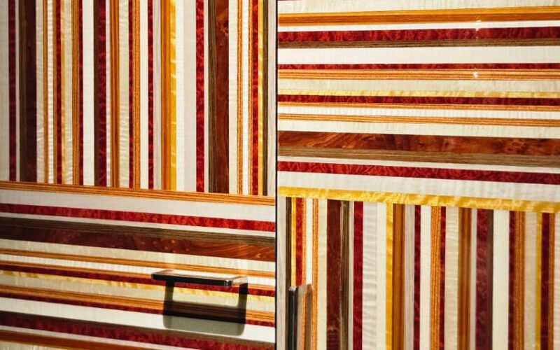 Marcenaria como obra de arte. Este armário usa lâminas de madeira em tons quentes (vermelho, amarelo, marrom) para criar um padrão gráfico de listras. A alternância entre horizontal e vertical, combinada com puxadores modernos, resulta em um design arrojado e luxuoso.