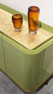 Design que une cor e textura. O aparador destaca-se pelas curvas suaves e pela laca brilhante em tom verde-oliva. O tampo rebaixado em madeira natural aquece a peça, enquanto os vidros âmbar texturizados adicionam um ponto de luz sofisticado.