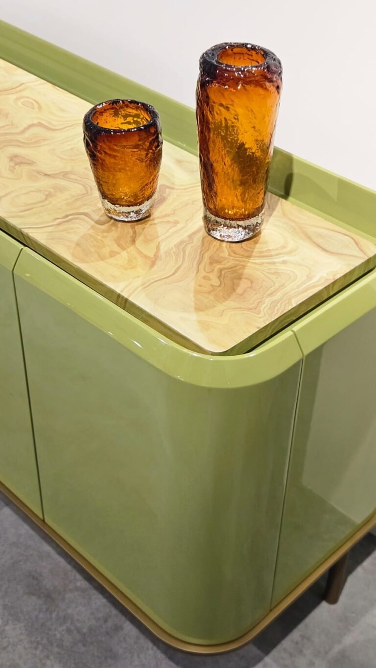 Design que une cor e textura. O aparador destaca-se pelas curvas suaves e pela laca brilhante em tom verde-oliva. O tampo rebaixado em madeira natural aquece a peça, enquanto os vidros âmbar texturizados adicionam um ponto de luz sofisticado.