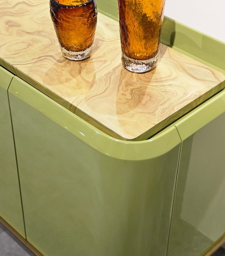 Design que une cor e textura. O aparador destaca-se pelas curvas suaves e pela laca brilhante em tom verde-oliva. O tampo rebaixado em madeira natural aquece a peça, enquanto os vidros âmbar texturizados adicionam um ponto de luz sofisticado.