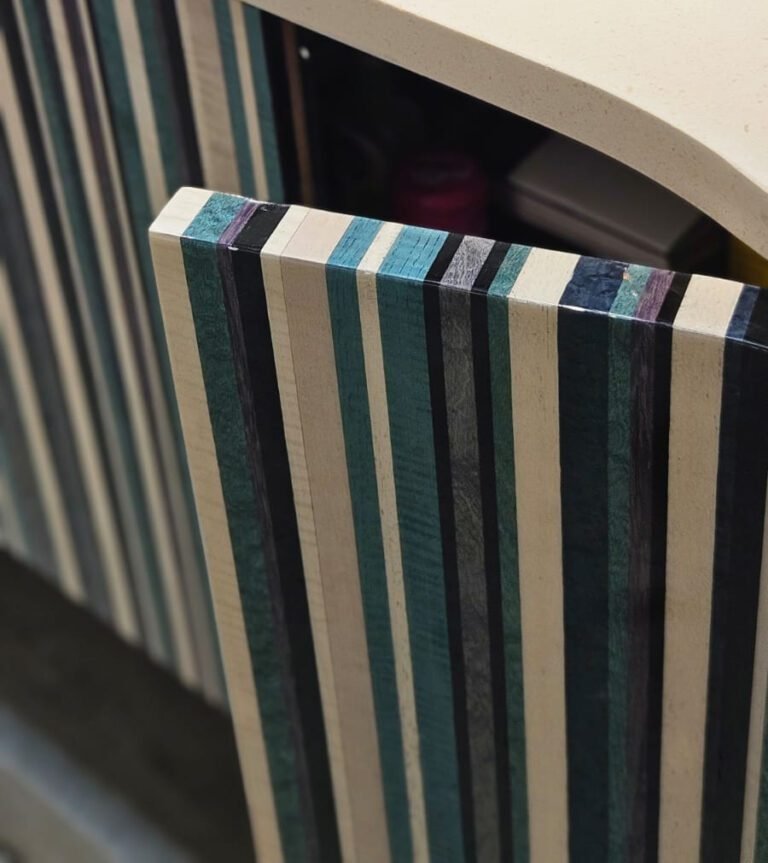 A marcenaria como arte. Este móvel revela sua sofisticação nas bordas e na porta curva, feitas de madeira laminada (multilaminado). As listras em tons de azul, verde, preto e natural criam um padrão gráfico vibrante. Um design que celebra o material.