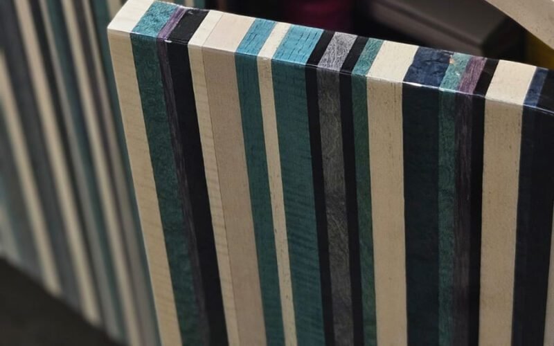 A marcenaria como arte. Este móvel revela sua sofisticação nas bordas e na porta curva, feitas de madeira laminada (multilaminado). As listras em tons de azul, verde, preto e natural criam um padrão gráfico vibrante. Um design que celebra o material.