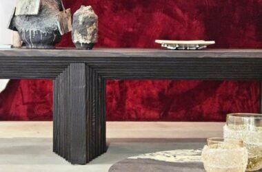 Luxo dramático focado em texturas. O veludo vermelho profundo da parede serve de fundo para o aparador em madeira ebanizada e ranhurada. A mesa de centro em pedra e os vasos esculturais adicionam camadas de sofisticação e uma sensação tátil ao ambiente.