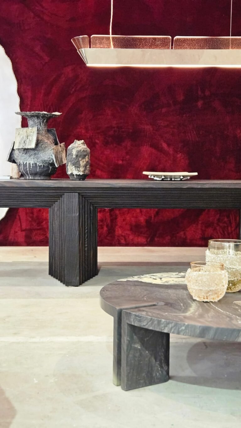 Luxo dramático focado em texturas. O veludo vermelho profundo da parede serve de fundo para o aparador em madeira ebanizada e ranhurada. A mesa de centro em pedra e os vasos esculturais adicionam camadas de sofisticação e uma sensação tátil ao ambiente.