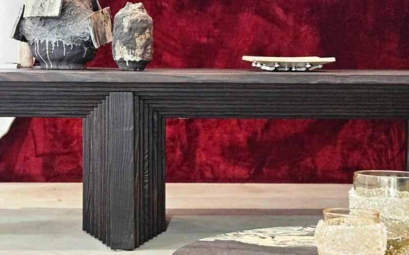 Luxo dramático focado em texturas. O veludo vermelho profundo da parede serve de fundo para o aparador em madeira ebanizada e ranhurada. A mesa de centro em pedra e os vasos esculturais adicionam camadas de sofisticação e uma sensação tátil ao ambiente.