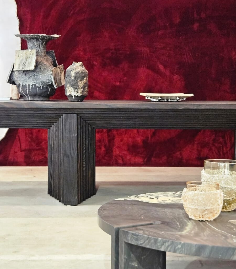 Luxo dramático focado em texturas. O veludo vermelho profundo da parede serve de fundo para o aparador em madeira ebanizada e ranhurada. A mesa de centro em pedra e os vasos esculturais adicionam camadas de sofisticação e uma sensação tátil ao ambiente.