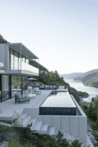 Piscina de alvenaria com borda infinita revestida em pedra escura, integrada a deck de madeira e vista para paisagem natural. Projeto de luxo ProjeFácil.
