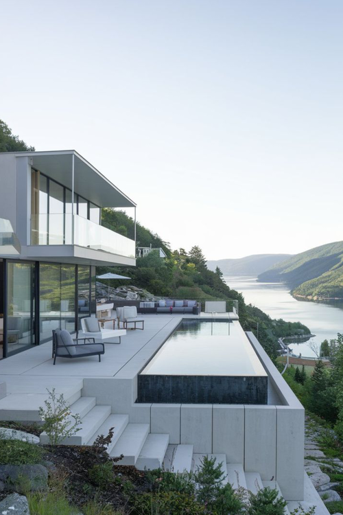 Piscina de alvenaria com borda infinita revestida em pedra escura, integrada a deck de madeira e vista para paisagem natural. Projeto de luxo ProjeFácil.