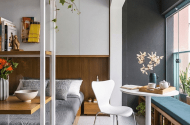 Visão geral de apartamento studio planejado integrando quarto com marcenaria amadeirada, cabeceira iluminada, cozinha compacta azul-petróleo e mesa de jantar redonda de mármore. Exemplo de como ampliar ambientes pequenos com design inteligente.