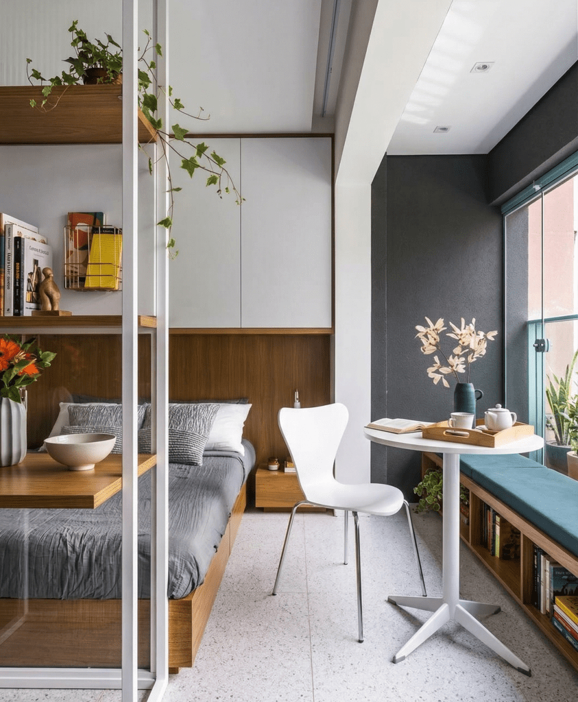 Visão geral de apartamento studio planejado integrando quarto com marcenaria amadeirada, cabeceira iluminada, cozinha compacta azul-petróleo e mesa de jantar redonda de mármore. Exemplo de como ampliar ambientes pequenos com design inteligente.